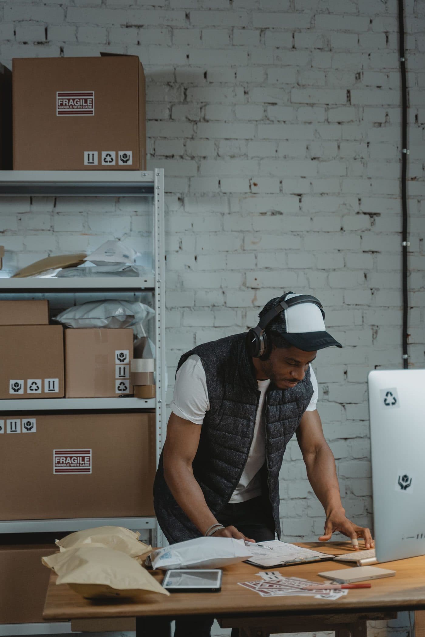 A man packing orders at a workbench beside his computer, boxes stacked on the shelves behind